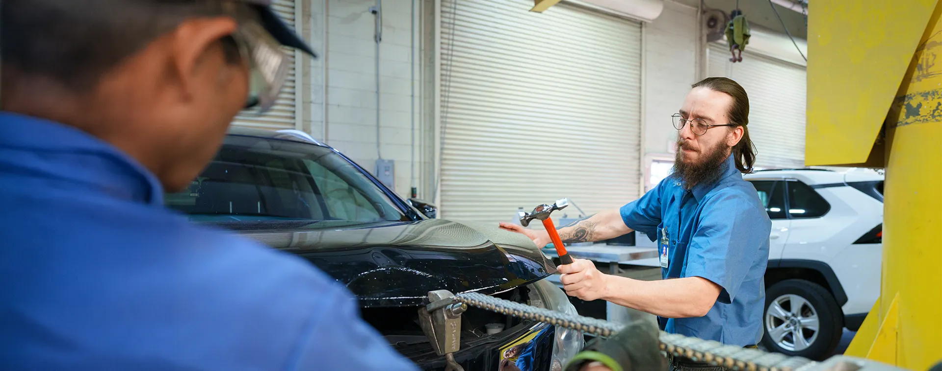 Student uses a body repair hammer on a vehicle panel in an automotive collision repair workshop.