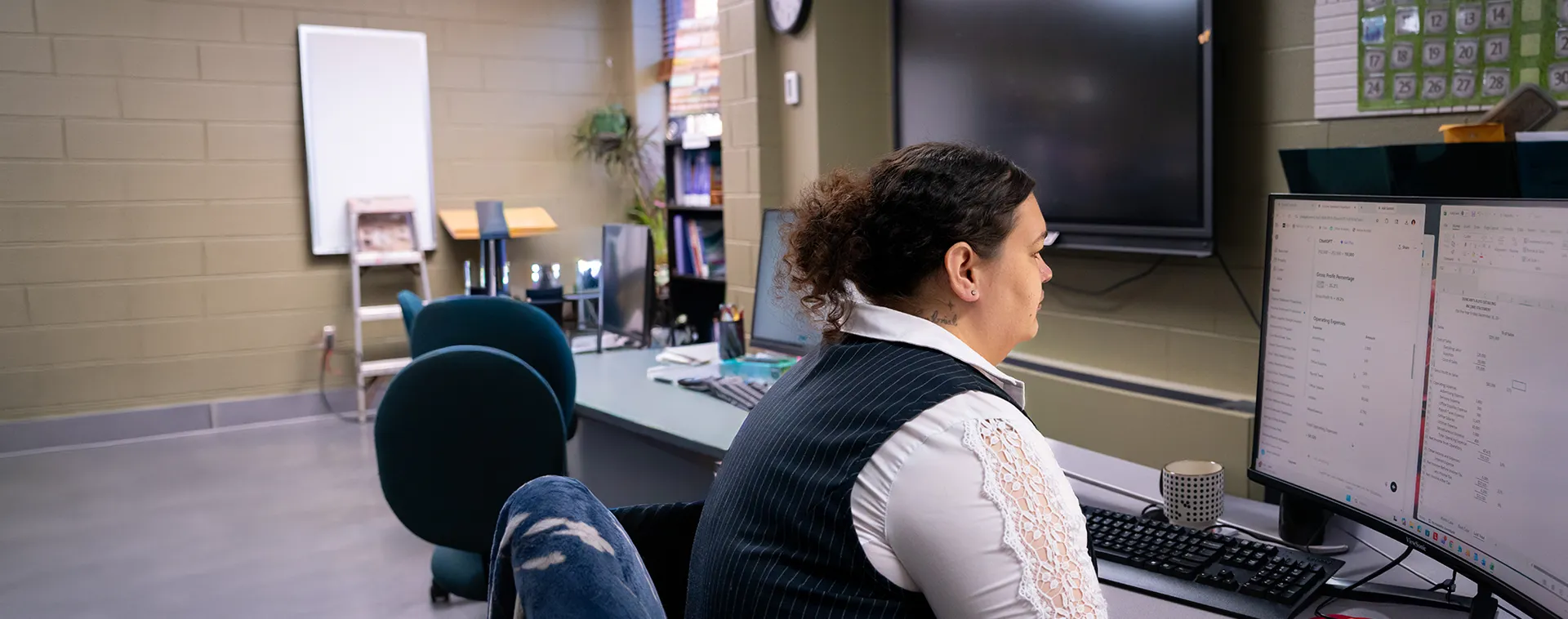 Person seated at desk using computer with dual monitors in classroom office, chairs, whiteboard, and large screen in background