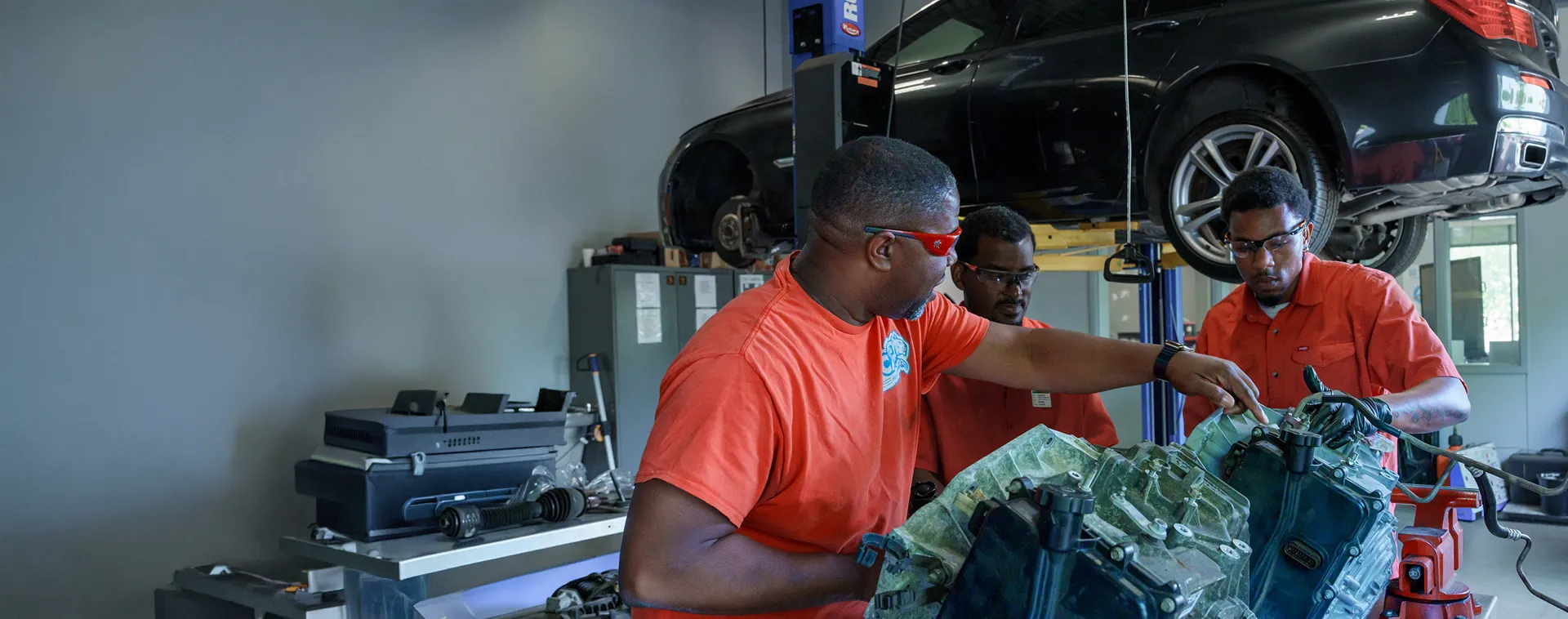 Students and instructor inspect an engine component in an automotive lab with a vehicle raised on a lift.