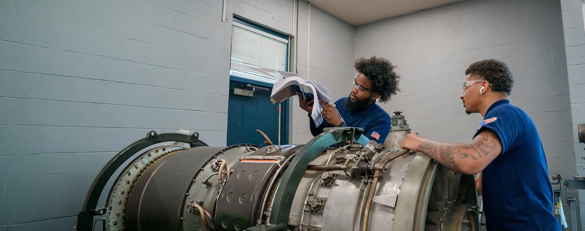 Two students review a manual beside a jet engine assembly in an aviation maintenance lab.