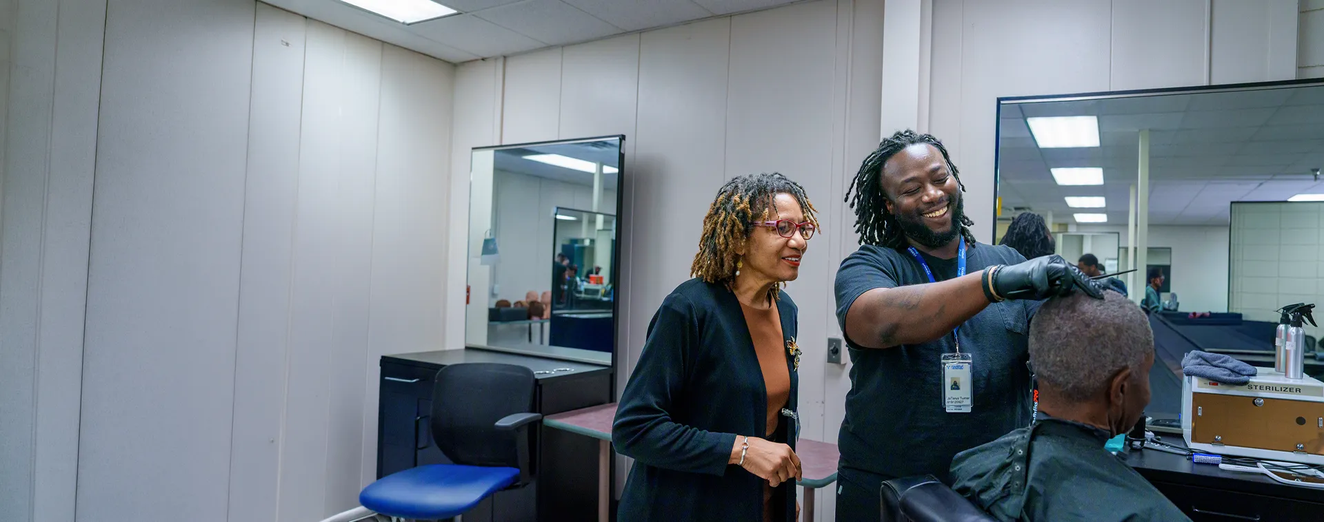 Barbering instructor observes a student cutting a client's hair in a training salon with mirrors and workstations.