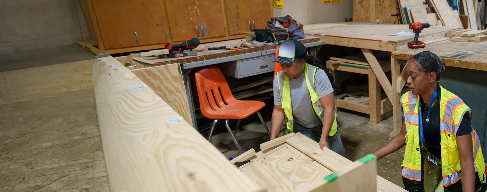 Students wearing safety vests assemble a plywood wall section in a building construction workshop.