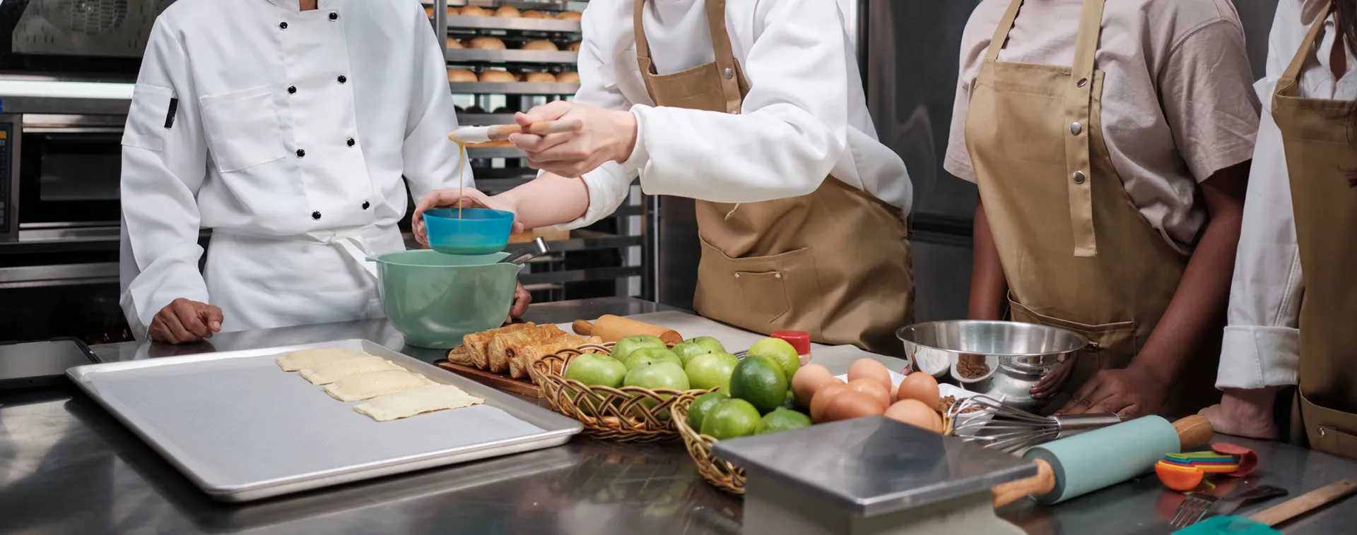 Students and instructor prepare pastry dough and ingredients at a stainless steel worktable in a commercial kitchen.