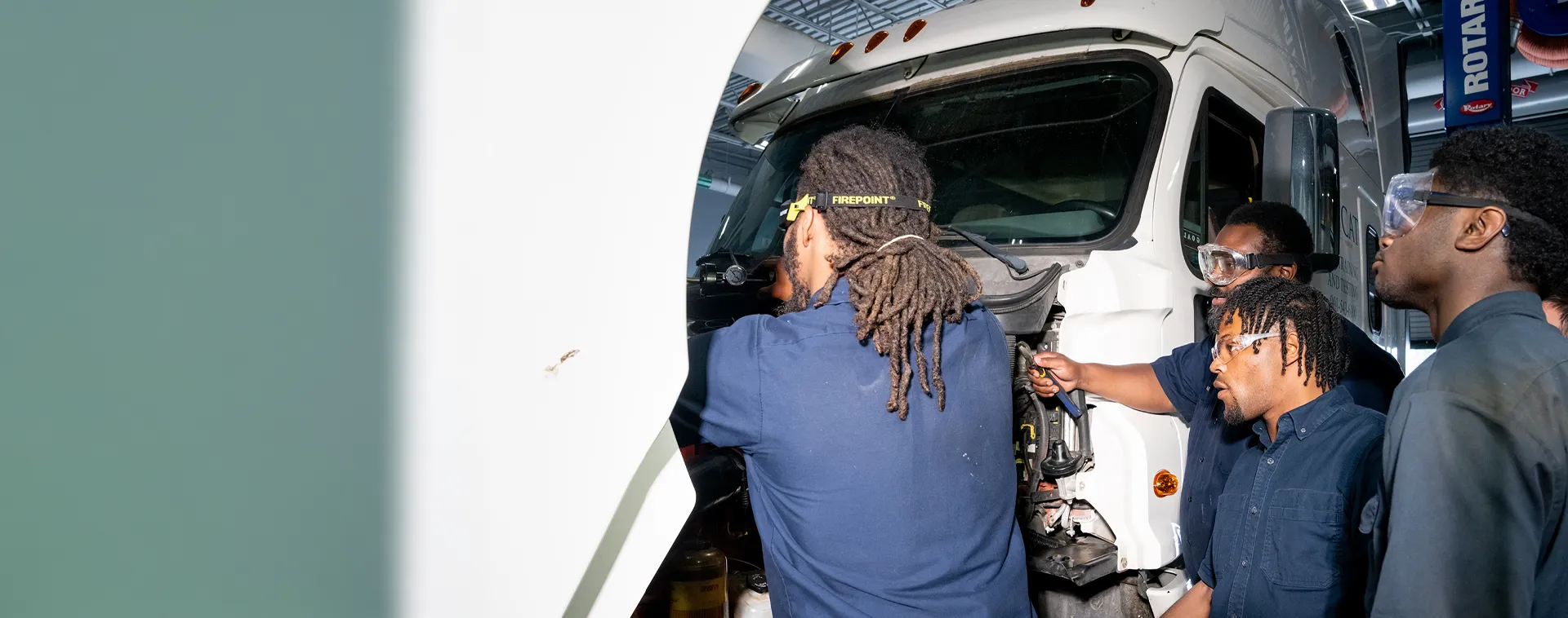 Students inspect components under the hood of a semi-truck in a diesel technology training shop.