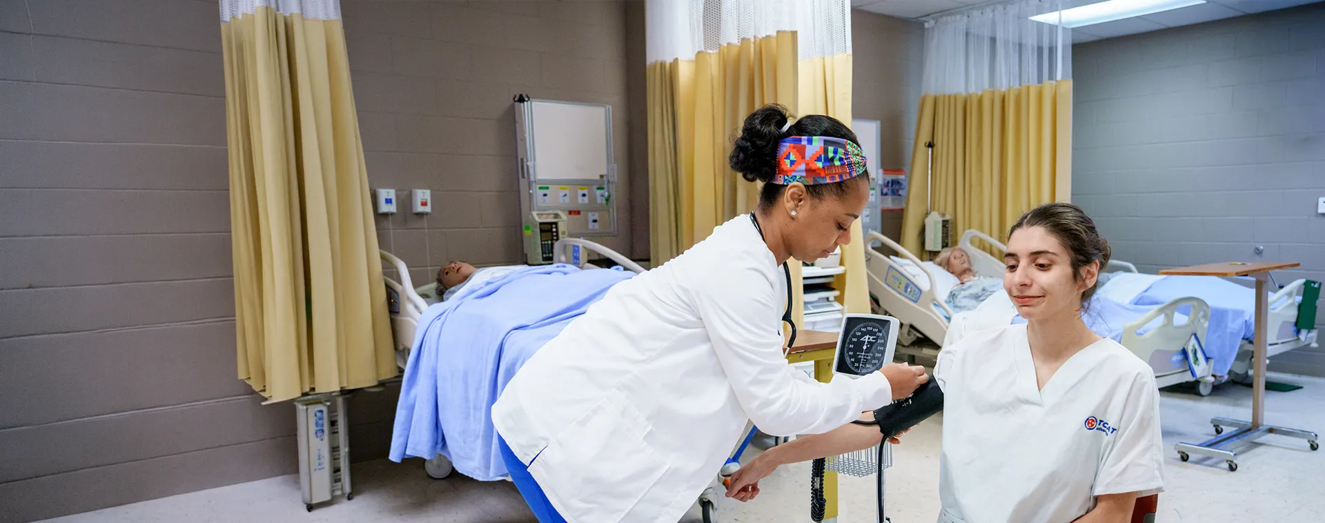 Student checks blood pressure on another student in a nursing lab with hospital beds and patient simulators in the background.