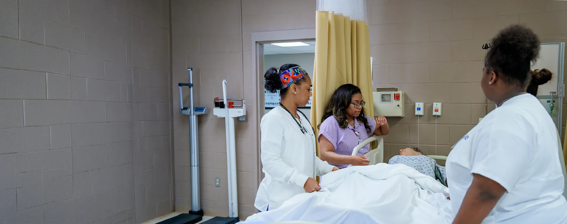 Nursing students practice patient care around a hospital bed in a clinical training lab.
