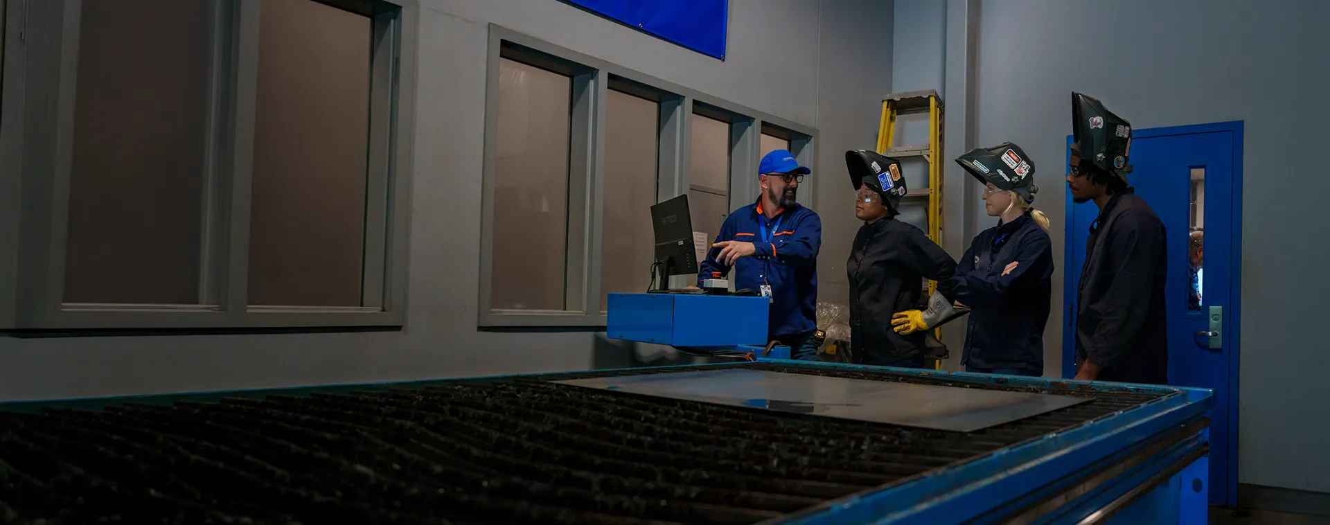Instructor reviews welding equipment controls with students wearing welding helmets in a welding lab.