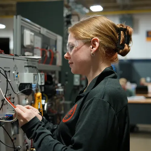 Student wearing safety glasses connects wiring on an HVAC training control panel.
