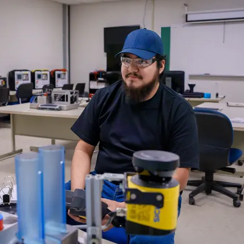Student wearing safety glasses sits beside industrial automation equipment in a technical training classroom.