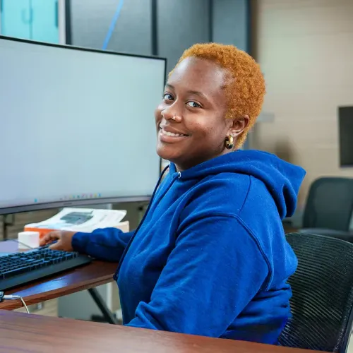 Student seated at a computer workstation looks toward the camera in a computer design lab.