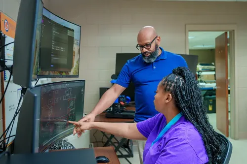 Instructor and student review architectural plans on dual monitors in a computer-aided design lab.