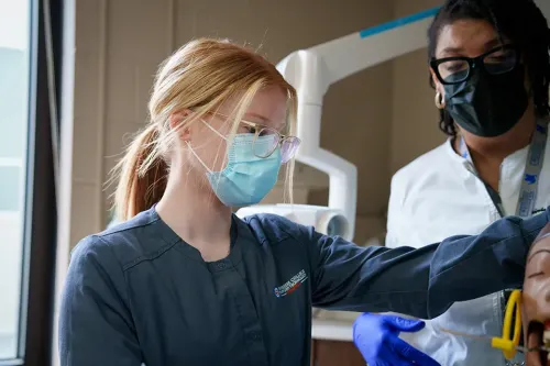 Student in scrubs and mask practices dental assisting skills with an instructor in a clinical lab.