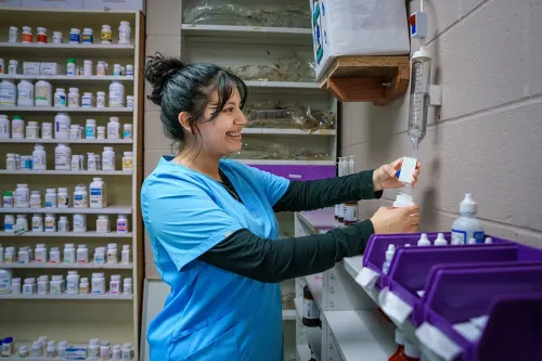 Student in scrubs measures medication from a dispensing device with pharmacy shelves in the background.